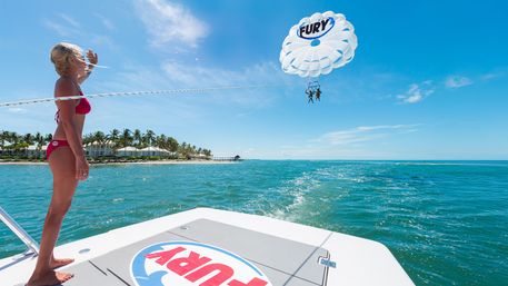 Person in a red bikini standing on a boat deck shielding their eyes while a white parasail lifts two riders over turquoise ocean near a palm-lined tropical shoreline under a bright blue sky.