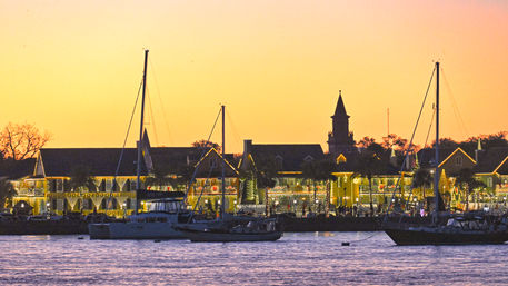 Golden-sky waterfront marina at sunset with anchored sailboats, festive holiday lights on harbor buildings and a silhouetted church steeple.