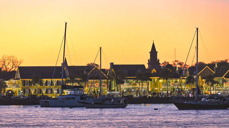 Golden-sky waterfront marina at sunset with anchored sailboats, festive holiday lights on harbor buildings and a silhouetted church steeple.