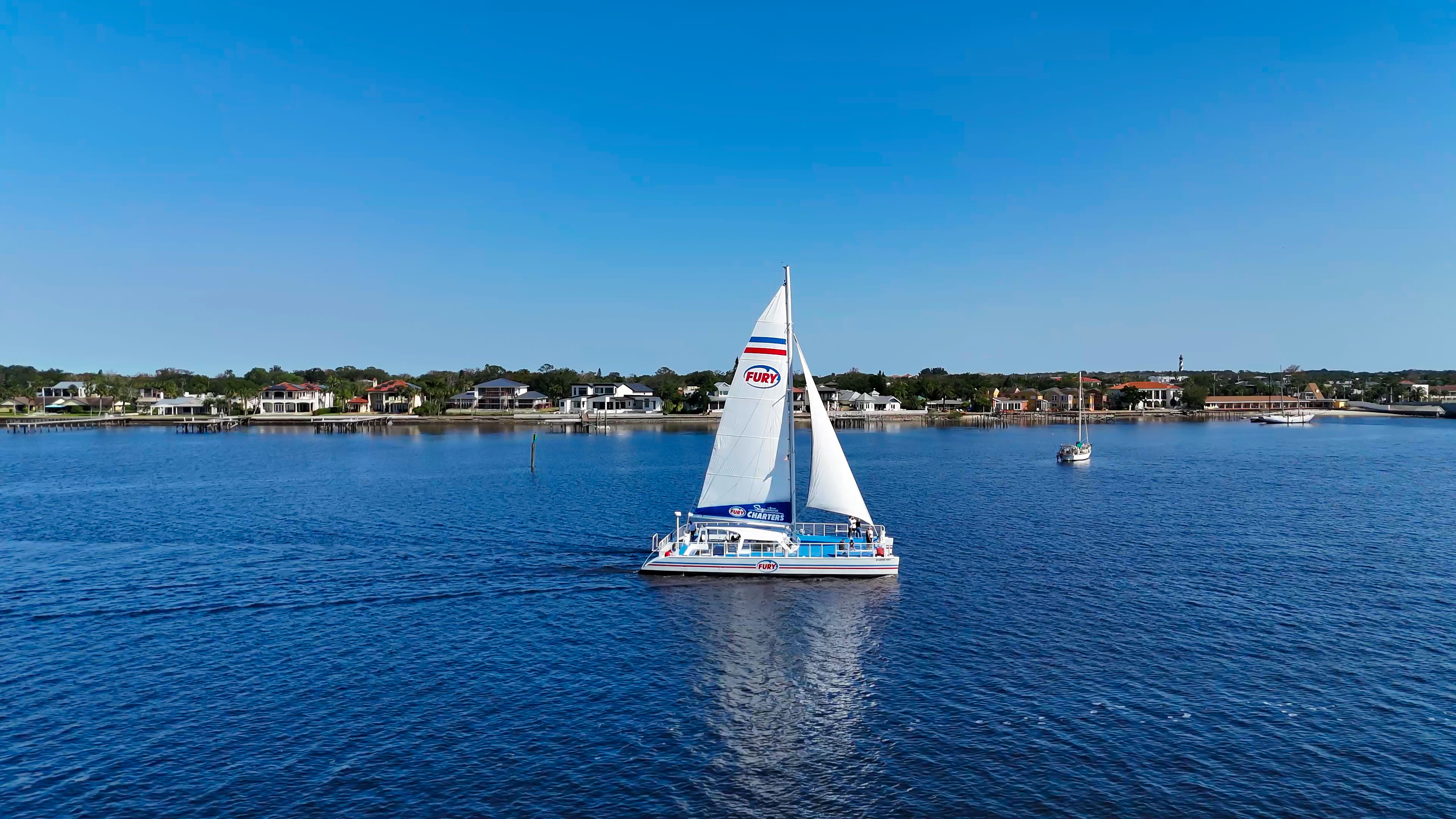 White sail catamaran cruising on a deep-blue coastal bay, sunlight reflecting on calm water under a clear sky with waterfront homes along the distant shoreline.