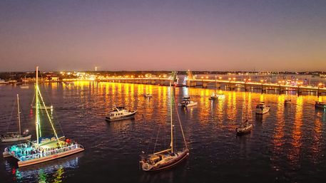 Twilight coastal harbor with sailboats and yachts anchored near an illuminated drawbridge, golden city lights and festive boat lights reflecting across calm water.