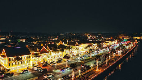 Nighttime waterfront street with palm trees and historic-style buildings twinkling in festive holiday lights, bustling with cars and pedestrians along a glowing coastal promenade.