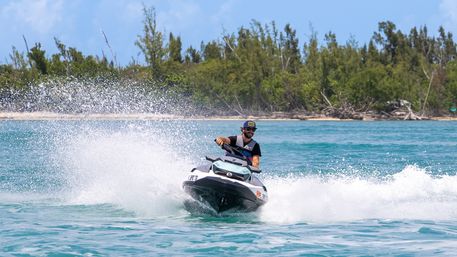 Person riding a jet ski across turquoise water, spray arcing by a tree-lined tropical island shoreline, upbeat watersports scene