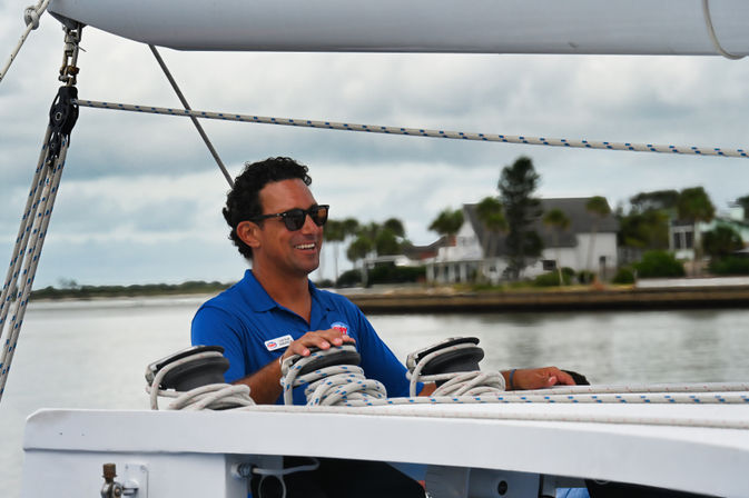 Smiling crew member in a blue polo and sunglasses adjusting coiled ropes and winches on a sailboat with waterfront homes and palm trees along a coastal shoreline under an overcast sky.