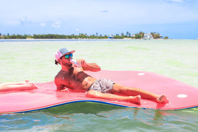 Man lounging on a pink float in shallow turquoise sea near a palm-lined island, wearing sunglasses and a trucker hat while sipping a canned drink.