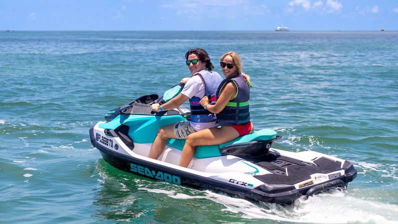 Two people in life vests smiling on a teal-and-white jet ski gliding across calm blue ocean waters on a sunny day