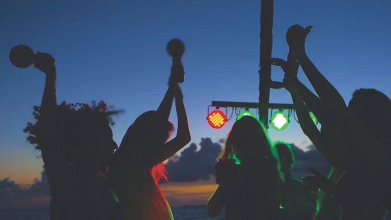 Silhouetted group dancing at a beach sunset party under red and green stage lights, hands raised and some holding handheld shakers — coastal outdoor nightlife vibe.