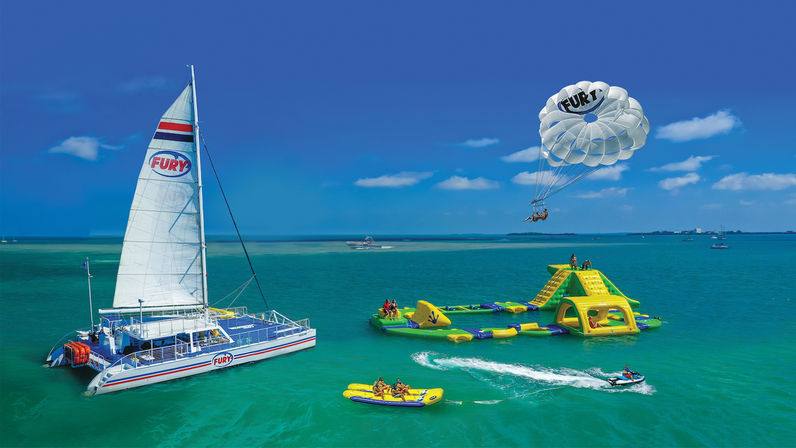 Sunny tropical scene with a white catamaran, colorful inflatable water park, parasail riders, banana boat and jet ski on turquoise sea
