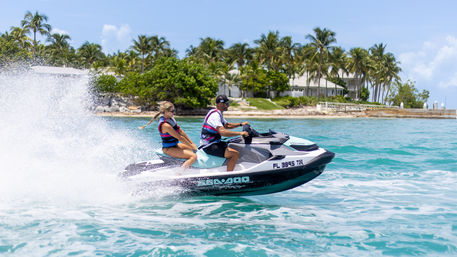 Two people in life vests riding a Sea‑Doo jet ski across turquoise tropical waters, spraying a big wake past a palm‑lined shoreline with beachfront homes.