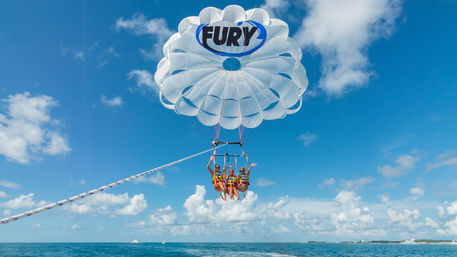 Three people in colorful life vests parasailing over turquoise ocean beneath a large white parachute against a bright blue sky with a distant coastline — tropical water sports scene.