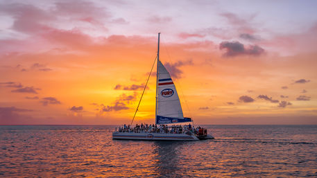 Catamaran with passengers sailing on a calm sea beneath a vivid orange-pink sunset sky, silhouetted against the horizon.