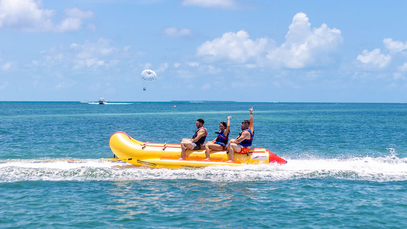 Three people in life jackets riding a bright yellow inflatable banana boat across turquoise coastal waters on a sunny day, with a parasail and boat visible on the horizon.