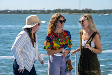 Three women laughing and chatting on a sunny boat deck by a blue bay, wearing sunglasses and summer outfits—one with a wide-brim hat and another in a colorful top holding a plate and cup.