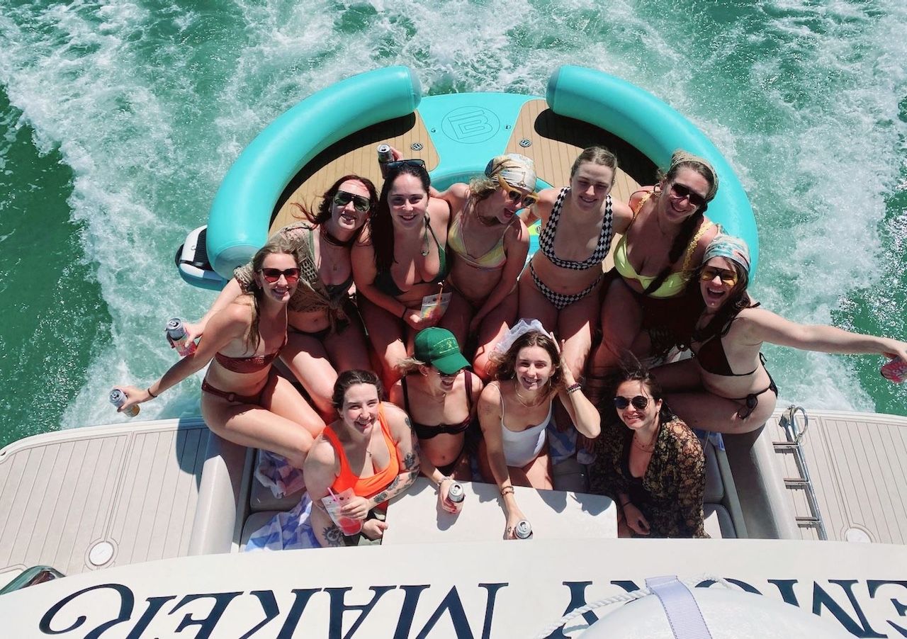 Overhead shot of a group of friends in swimsuits partying on the stern of a motorboat with a teal inflatable, holding drinks and smiling as white wake churns through green water on a sunny summer day.
