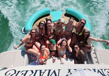 Overhead shot of a group of friends in swimsuits partying on the stern of a motorboat with a teal inflatable, holding drinks and smiling as white wake churns through green water on a sunny summer day.