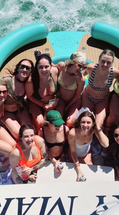 Overhead shot of a group of friends in swimsuits partying on the stern of a motorboat with a teal inflatable, holding drinks and smiling as white wake churns through green water on a sunny summer day.