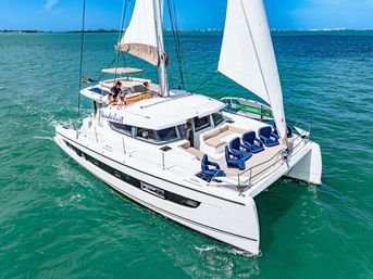 Luxury white catamaran yacht with raised sails and blue deck chairs cruising on turquoise water near a distant coastal skyline under a sunny sky
