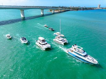 Aerial view of luxury yachts, catamarans and powerboats cruising sunlit turquoise coastal waters beneath a modern concrete bridge with a distant shoreline.