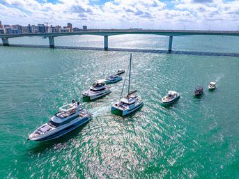 Aerial view of luxury yachts and small boats clustered in sparkling turquoise coastal water near a long concrete bridge and city skyline under a sunny, partly cloudy sky.