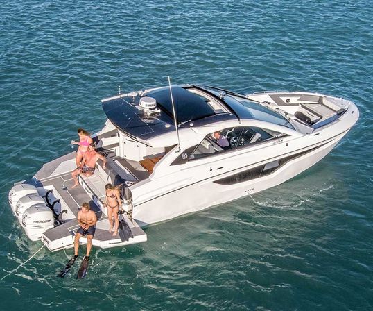 Aerial shot of a white luxury motorboat anchored in clear blue coastal water on a sunny day, four people on the swim platform gearing up to snorkel and sunbathe with triple outboard engines and spacious bow seating.