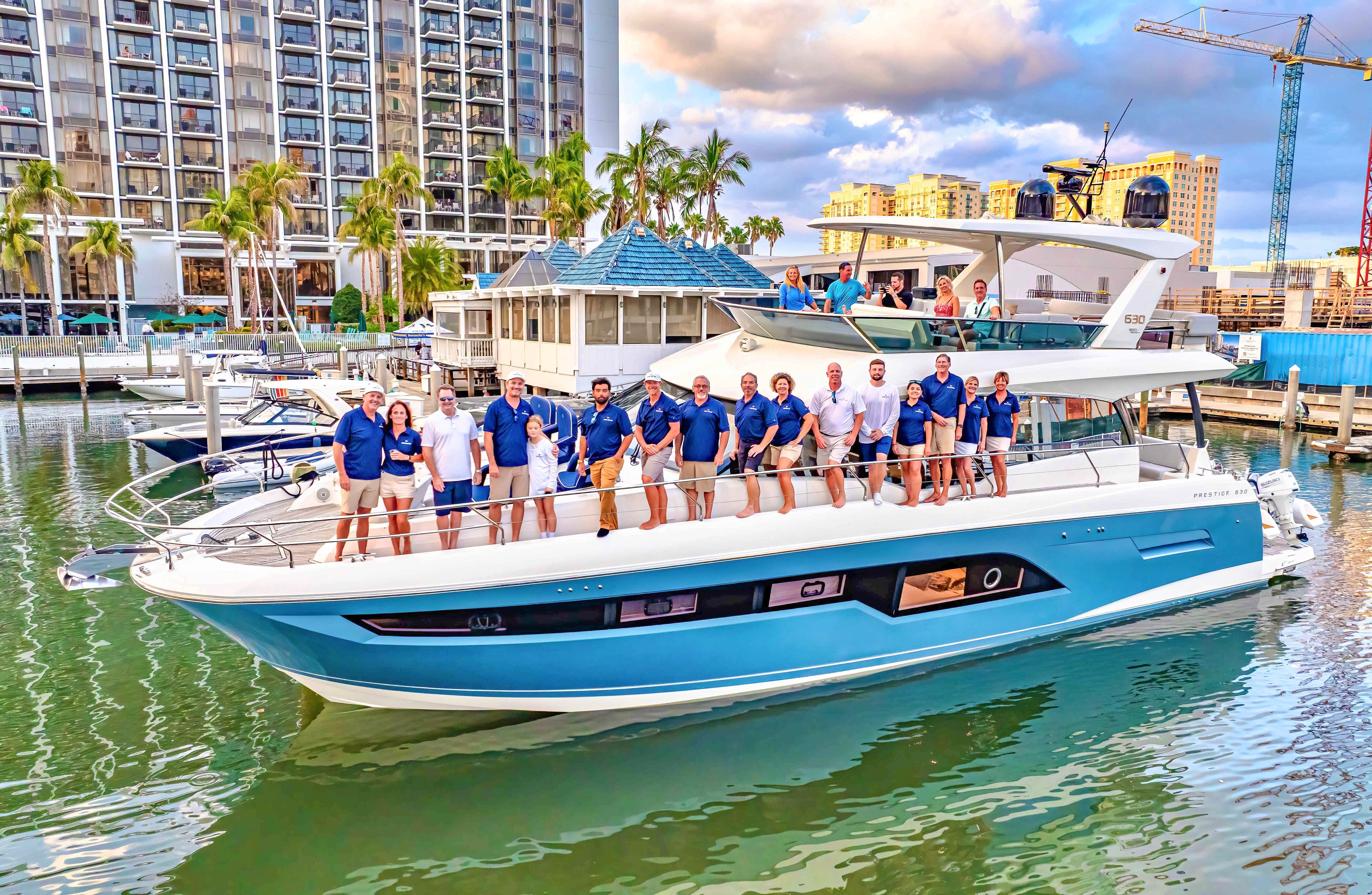 Luxury blue-and-white yacht at a palm-lined marina with a group of crew and guests posing on deck, waterfront high-rise hotel and boats in the background under a partly cloudy sky.