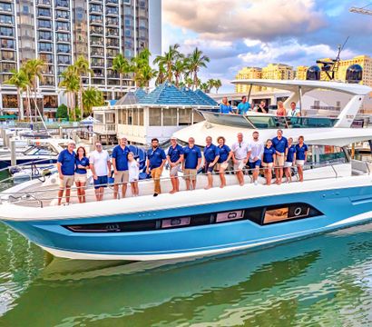 Luxury blue-and-white yacht at a palm-lined marina with a group of crew and guests posing on deck, waterfront high-rise hotel and boats in the background under a partly cloudy sky.
