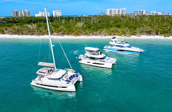 Aerial shot of three luxury boats — a white sailing catamaran and two motor yachts — floating in sunlit turquoise coastal water off a sandy beach with tree-lined shoreline and beachfront high-rise buildings.