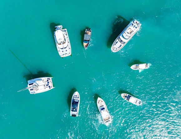 Aerial view of yachts and motorboats scattered in sunlit turquoise water, their white hulls, wakes, and shadows forming a striking pattern.
