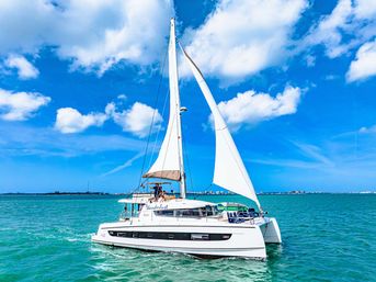 White sailing catamaran with full sails gliding on turquoise water under a bright blue sky with fluffy clouds and a distant coastal skyline.