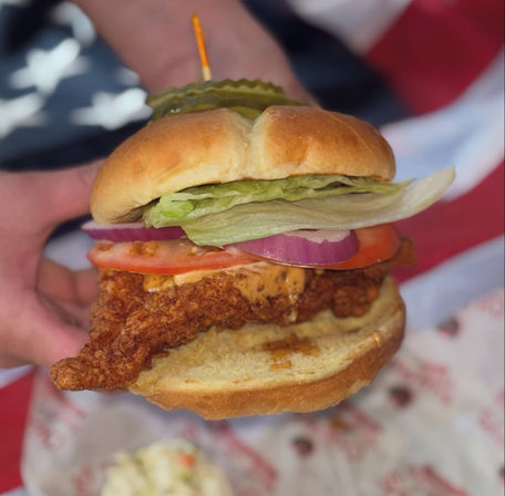 Close-up handheld crispy fried chicken sandwich with lettuce, tomato, red onion, pickles and creamy sauce on a toasted bun against a flag-themed picnic backdrop.