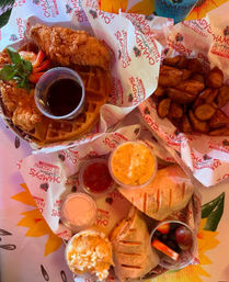 Crispy fried chicken on a waffle with syrup, roasted potato wedges, grilled sandwich halves, creamy mac and cheese, small side salads and a fruit cup — colorful Southern-style brunch spread.