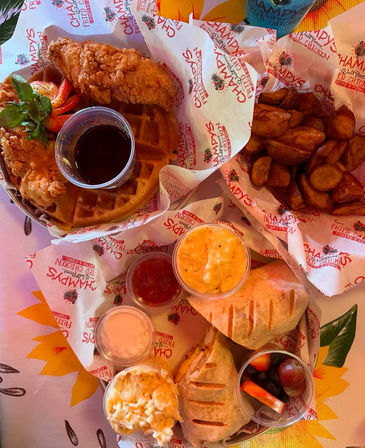 Crispy fried chicken on a waffle with syrup, roasted potato wedges, grilled sandwich halves, creamy mac and cheese, small side salads and a fruit cup — colorful Southern-style brunch spread.
