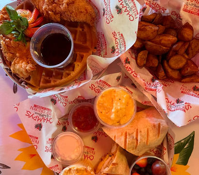 Crispy fried chicken on a waffle with syrup, roasted potato wedges, grilled sandwich halves, creamy mac and cheese, small side salads and a fruit cup — colorful Southern-style brunch spread.
