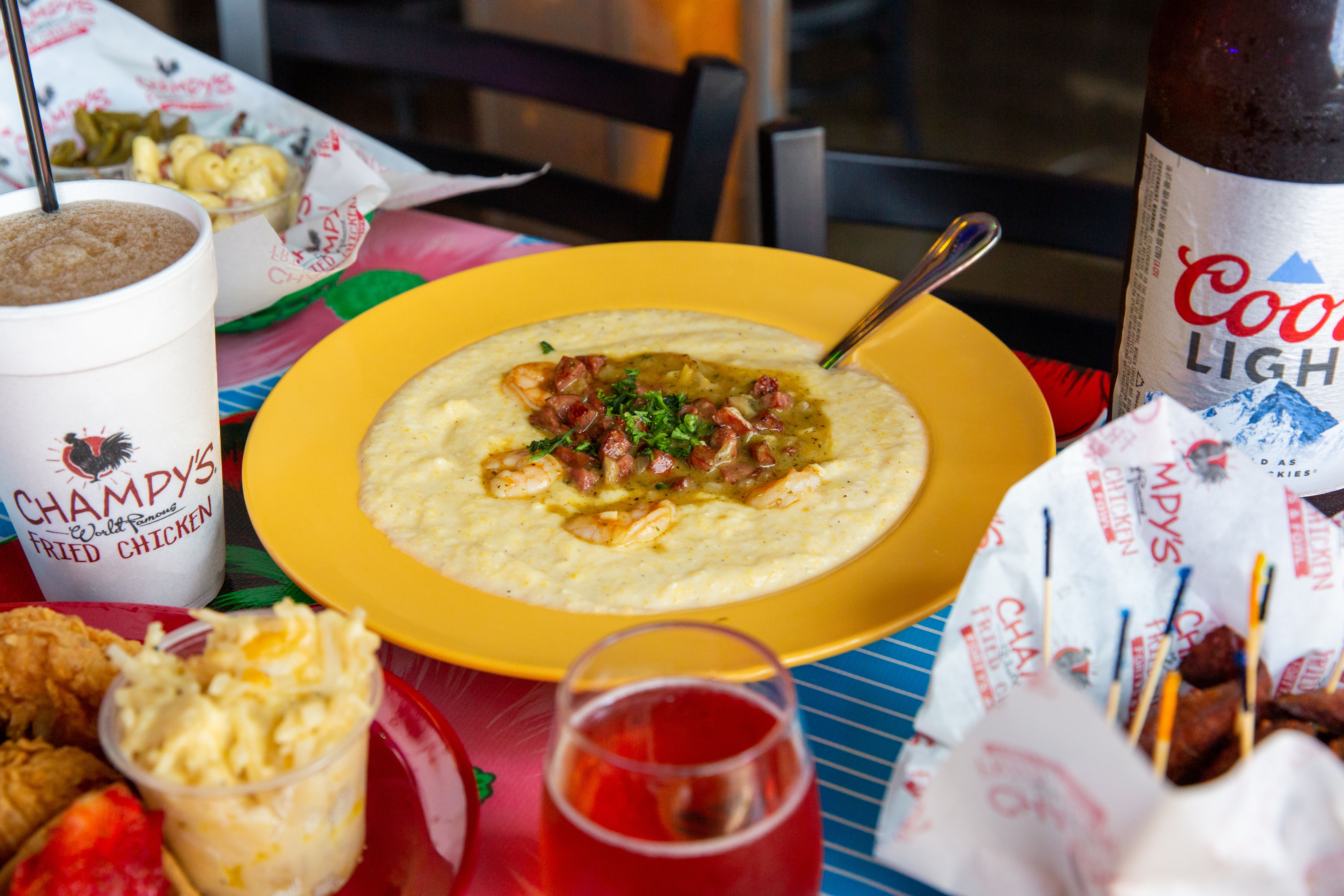 Hearty Southern shrimp and grits topped with shrimp, diced sausage and green gravy on a yellow plate, surrounded by fried chicken, macaroni salad, a soft drink cup and a beer bottle on a colorful restaurant table.