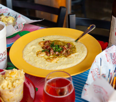 Hearty Southern shrimp and grits topped with shrimp, diced sausage and green gravy on a yellow plate, surrounded by fried chicken, macaroni salad, a soft drink cup and a beer bottle on a colorful restaurant table.
