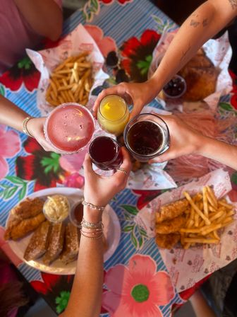 Overhead view of four hands toasting with a pink cocktail, orange juice, dark beer and red drink over a colorful floral tablecloth, surrounded by plates of fried chicken tenders, French fries and sliced toast — casual brunch with friends.