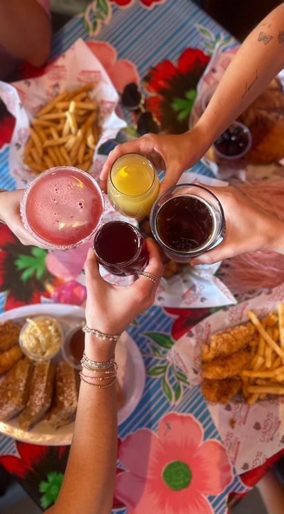 Overhead view of four hands toasting with a pink cocktail, orange juice, dark beer and red drink over a colorful floral tablecloth, surrounded by plates of fried chicken tenders, French fries and sliced toast — casual brunch with friends.