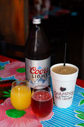 Beer bottle, two small glasses with orange and red drinks, and a styrofoam cup with a frozen drink and straw on a bright floral tablecloth at a casual fried-chicken restaurant.