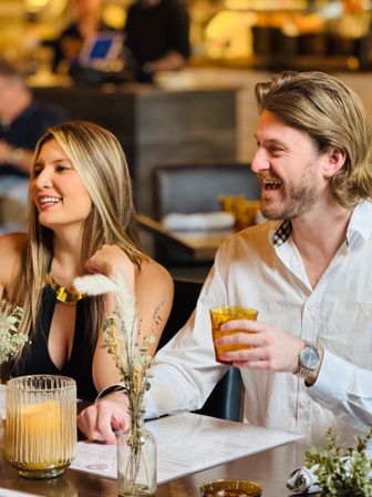 Smiling couple enjoying drinks at a cozy restaurant table with amber glassware, menus, and a dried floral centerpiece.