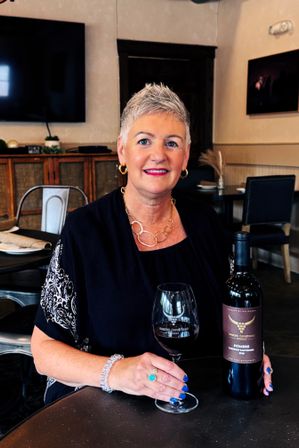Woman with short gray hair in a black top holding a glass of red wine and a bottle at a cozy winery tasting room table, wearing gold hoop earrings, circular gold necklace and turquoise ring — rustic modern wine tasting scene
