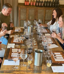 Group enjoying a wine tasting in a rustic winery tasting room — wooden table set with wine flights, tasting notes, spittoons and small snack cups.