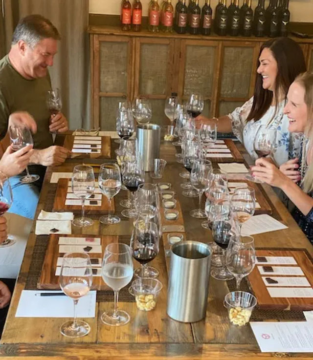 Group enjoying a wine tasting in a rustic winery tasting room — wooden table set with wine flights, tasting notes, spittoons and small snack cups.