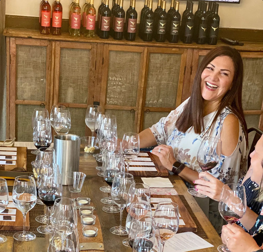 Smiling woman laughing during a winery tasting, seated at a rustic wooden table lined with rows of wine glasses, tasting notes and small snacks, with wine bottles on a shelf behind.