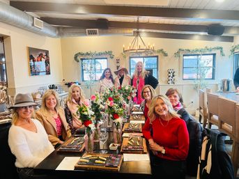 Group of women enjoying a wine-tasting brunch around a long wooden table set with cheese boards, wine glasses and colorful floral centerpieces in a bright rustic tasting room with a chandelier and greenery.