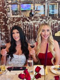 Two smiling friends toasting glasses of red wine at a rustic wood-paneled wine bar during a wine tasting, table set with red roses, a cheese plate and a tall burgundy candle