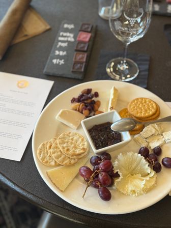 Cheese plate with crackers, assorted cheeses, red grapes, fruit preserve and nuts on a white plate beside a wine glass at a winery tasting.