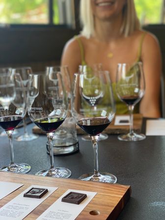 Sunlit winery tasting room table with a red wine flight in stemmed glasses and chocolate squares on a wooden tasting board, smiling guest blurred in the background.
