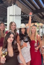 Group of smiling women in summer dresses and a cowboy hat holding wine glasses on an outdoor patio under a wooden pergola, one woman raising her hand with text overlay "where the hell is my husband?"