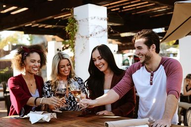 Four friends smiling and clinking white wine glasses on a sunlit outdoor restaurant patio around a wooden table.