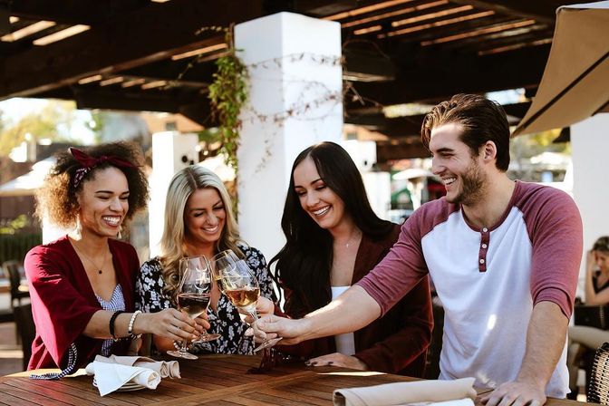 Four friends smiling and clinking white wine glasses on a sunlit outdoor restaurant patio around a wooden table.
